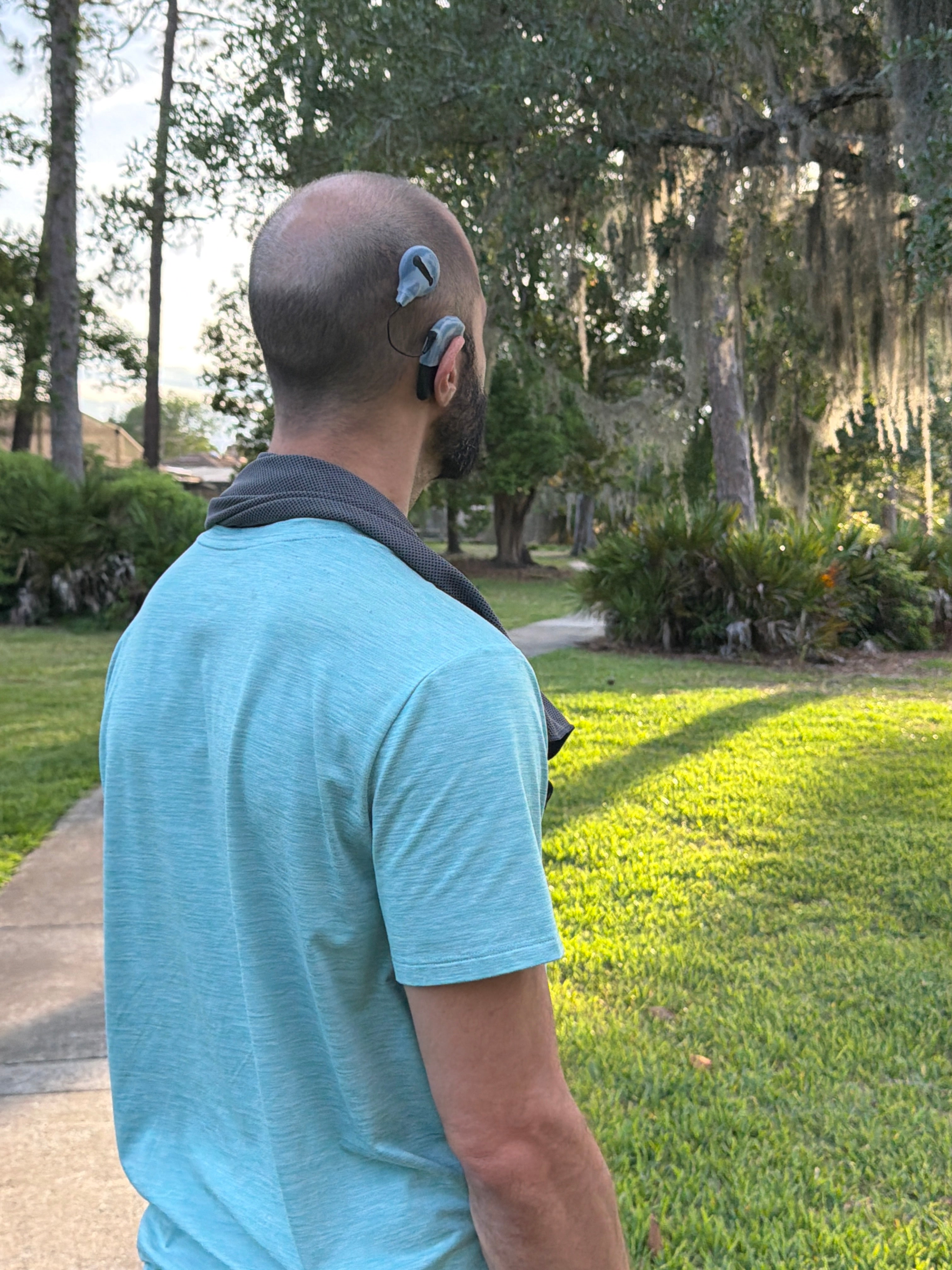 Person wearing a light blue shirt and a hearing implant with a protective silicone sleeve, standing outdoors with greenery and water in the background.